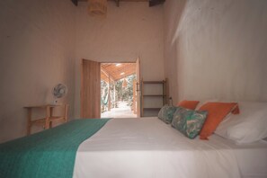 A bedroom with a bed, a fan, a wooden nightstand, and a view through an open door.