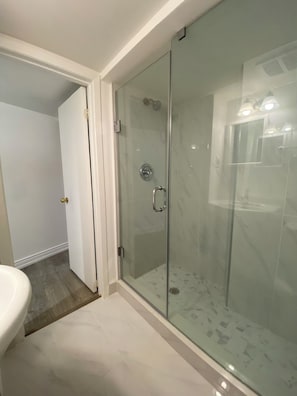 Une salle de bain moderne avec une douche à l’italienne en verre, un lavabo blanc et une porte donnant sur une autre pièce.