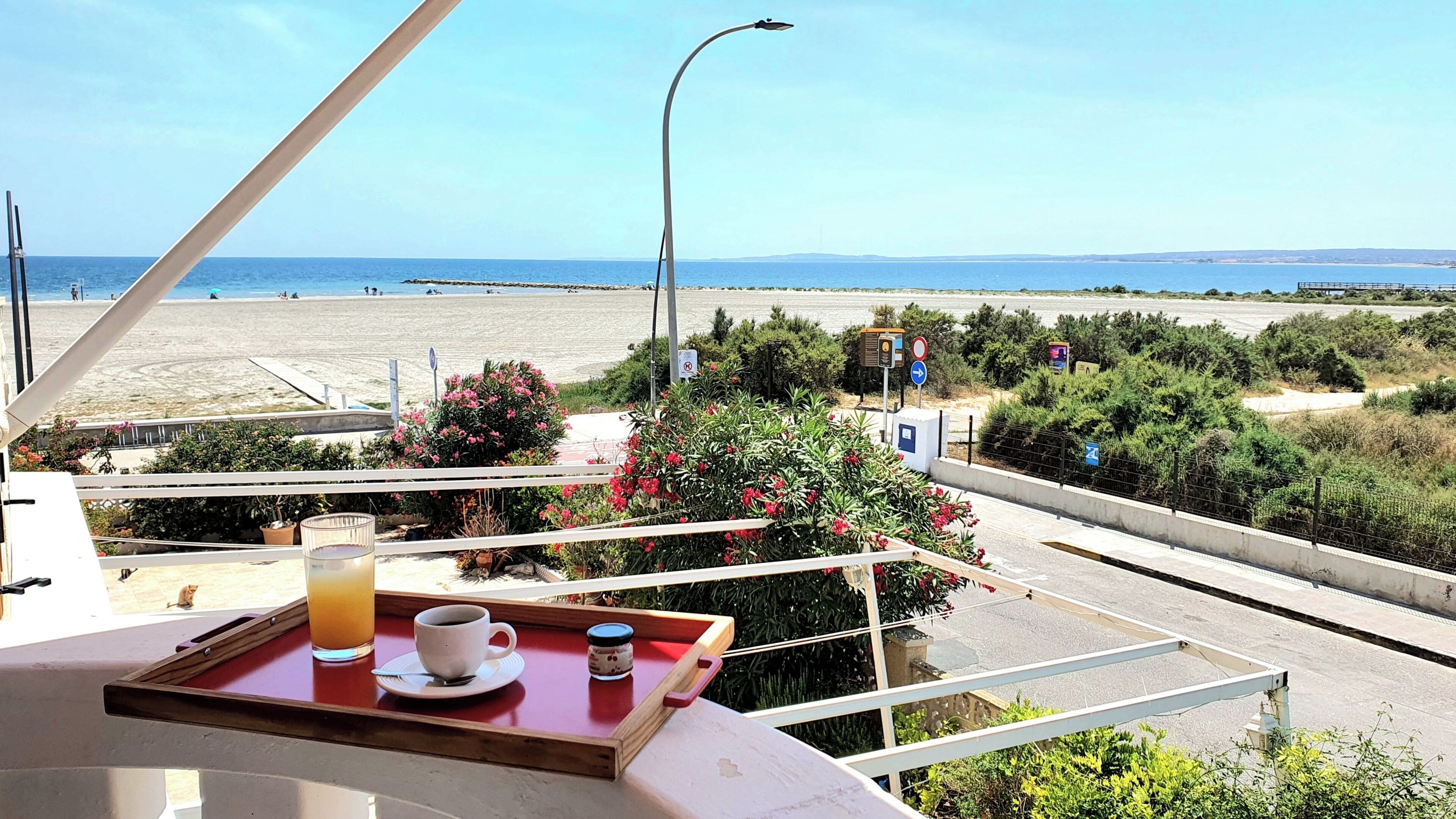 A balcony with a table, a cup, and a glass of juice, overlooking a beach and the sea.