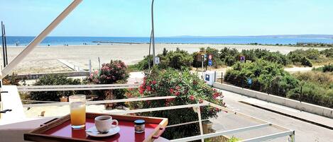 A balcony with a table, a cup, and a glass of juice, overlooking a beach and the sea.