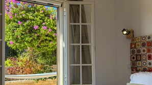 Ein Schlafzimmer mit großem Fenster, einem Bett mit weißer Bettwäsche, einer Wandleuchte und einem gemusterten Kopfteil.