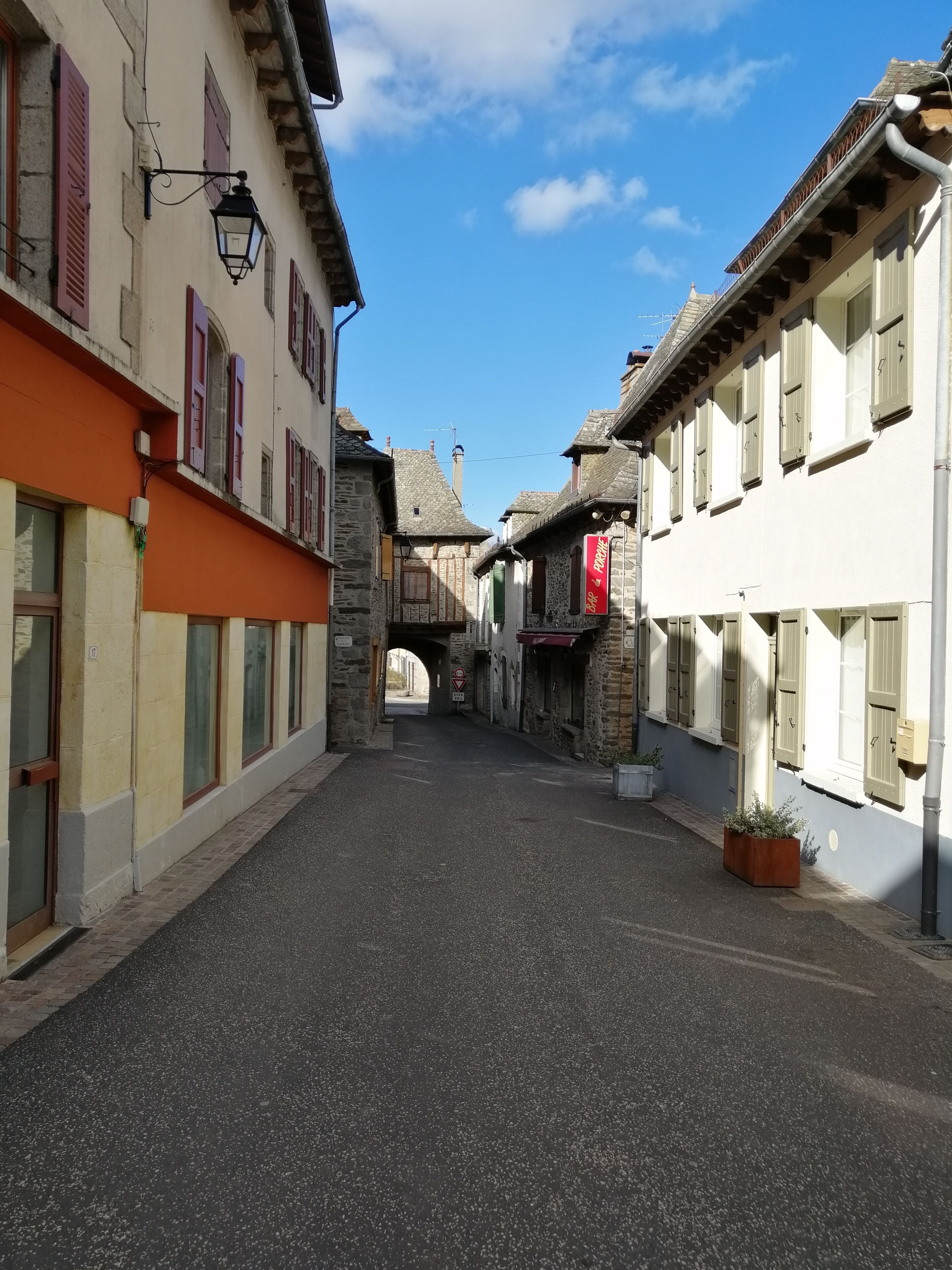 A narrow street with buildings on either side, a stone bridge in the distance, and a clear blue sky.