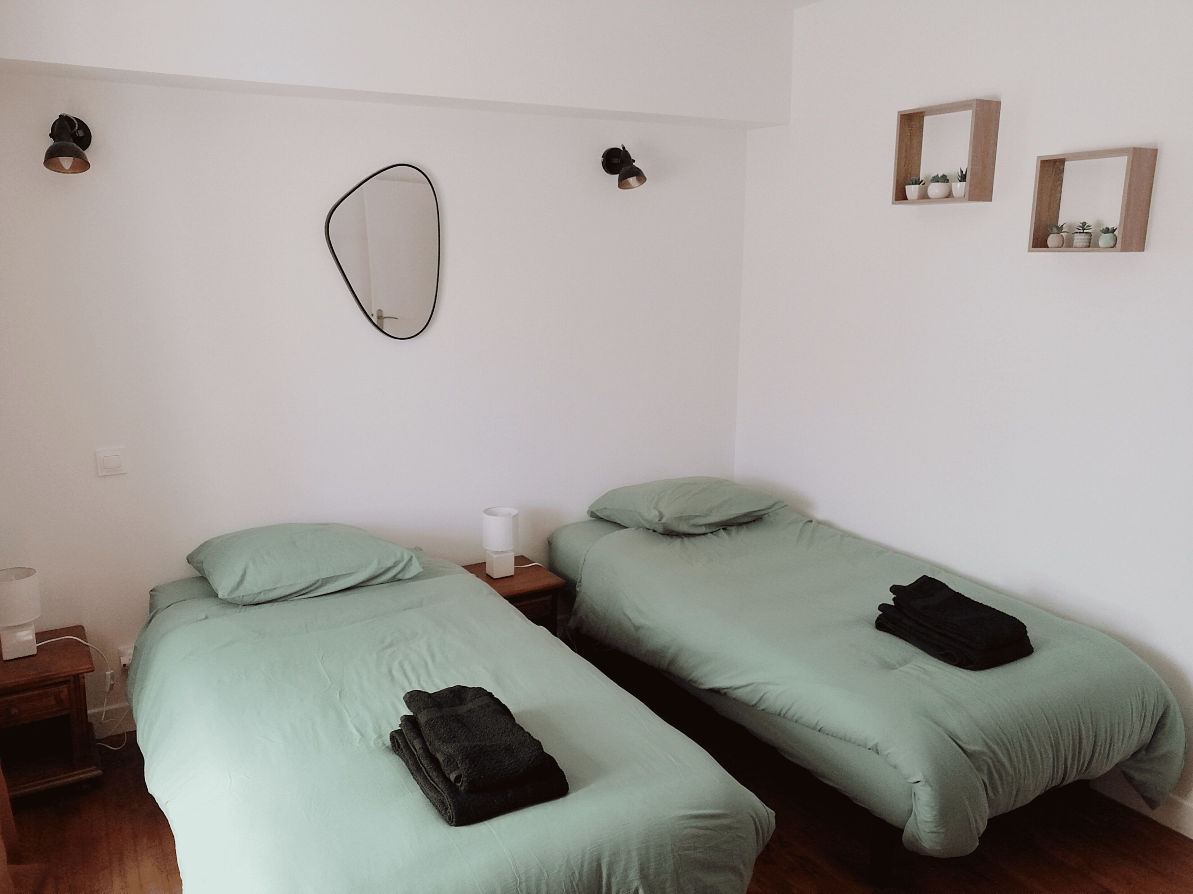 Una habitación con dos camas individuales, una mesita de noche con lámpara, un espejo en la pared y dos luces de pared.
