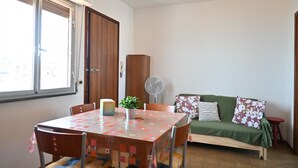 A dining area with a table, chairs, a fan, a sofa, and a window with blinds. - SE278 – Senigallia, seafront two-bedroom apartment with balconies and parking space (Senigallia)
