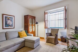 A living room with a sofa, a wicker chair, a cabinet, a framed picture, and a window with bars. - Renovated Historic Apartment with an Enclosed Terrace (Lisboa)