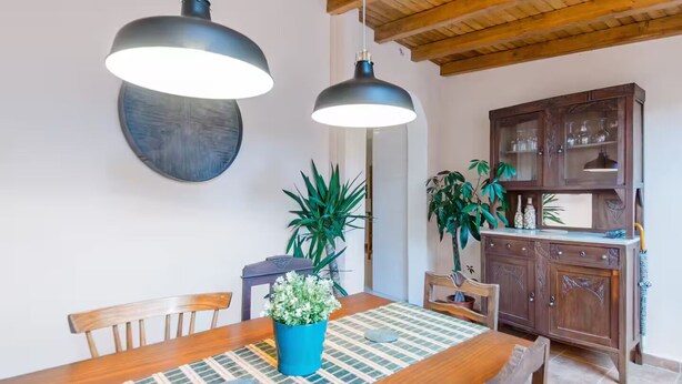 Un coin repas comprenant une table en bois, des chaises, deux lampes suspendues, une plante en pot et un buffet en bois contenant de la verrerie.
