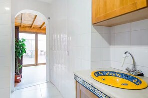 A modern kitchen with white tiles, a yellow sink with a blue pattern, wooden cabinets, and a potted plant. - Renovated Historic Apartment with an Enclosed Terrace (Lisboa)