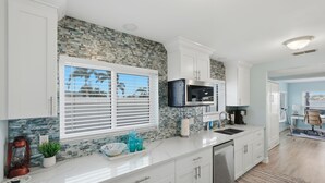 Una cocina con armarios blancos, protector de piedra salpicada, una ventana con persianas, un microondas y un fregadero.