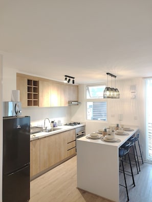 A modern kitchen with wooden cabinets, a black refrigerator, a gas stove, and a dining area with a white island and bar stools. - Oceana Suites Mirador Peñuelas II (Coquimbo)