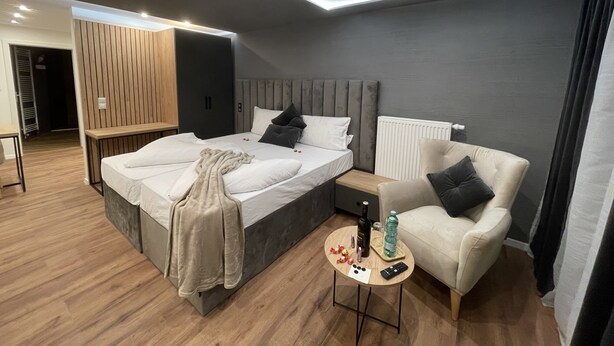 A modern bedroom with a bed, bedside table, chair, and a small table with a bottle and remote. - Apartmenthaus Zeuthen 3 (Zeuthen)