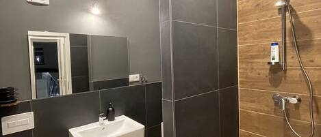 A modern bathroom with a white sink, a mirror, a shower, and dark tiled walls.