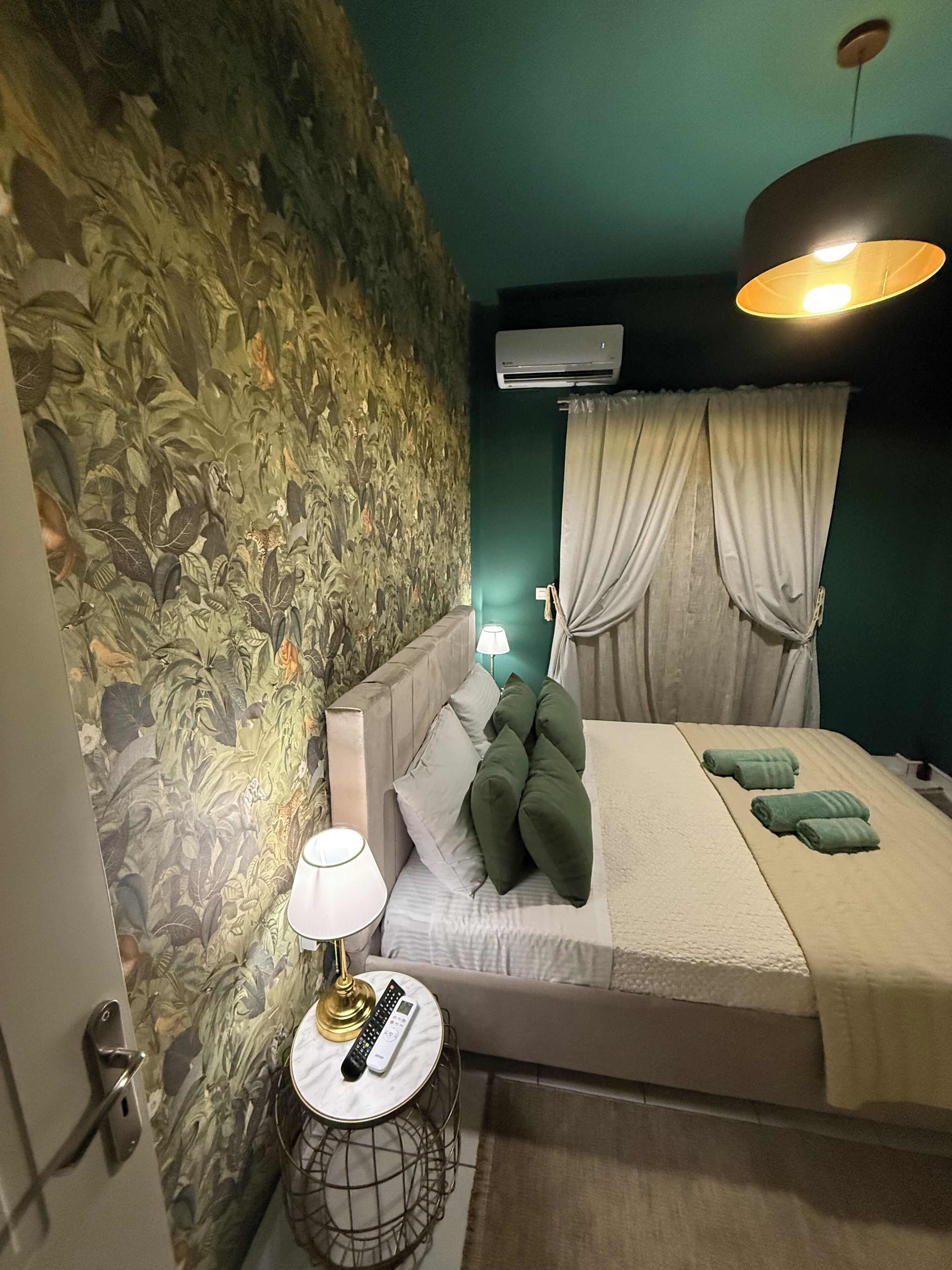 A bedroom with a bed, a round side table with a lamp, a remote control, and a wall-mounted air conditioner.