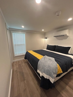 A bedroom with a bed, a window with blinds, an air conditioning unit, and a wooden floor. - Harbor Light | Modern Tiny Home with River Charm (Vicksburg)
