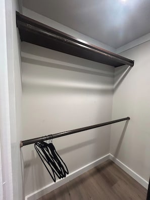 A room with a white wall, a wooden floor, and a metal shelf unit with a hanging rack. - Harbor Light | Modern Tiny Home with River Charm (Vicksburg)