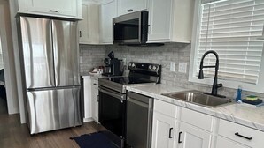 A modern kitchen with white cabinetry, stainless steel appliances, a black stove, and a window with blinds. - Harbor Light | Modern Tiny Home with River Charm (Vicksburg)