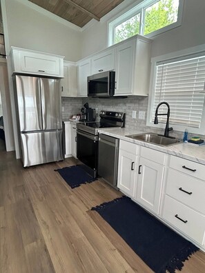 A modern kitchen with white cabinetry, stainless steel appliances, a black stove, and a window with blinds. - Harbor Light | Modern Tiny Home with River Charm (Vicksburg)