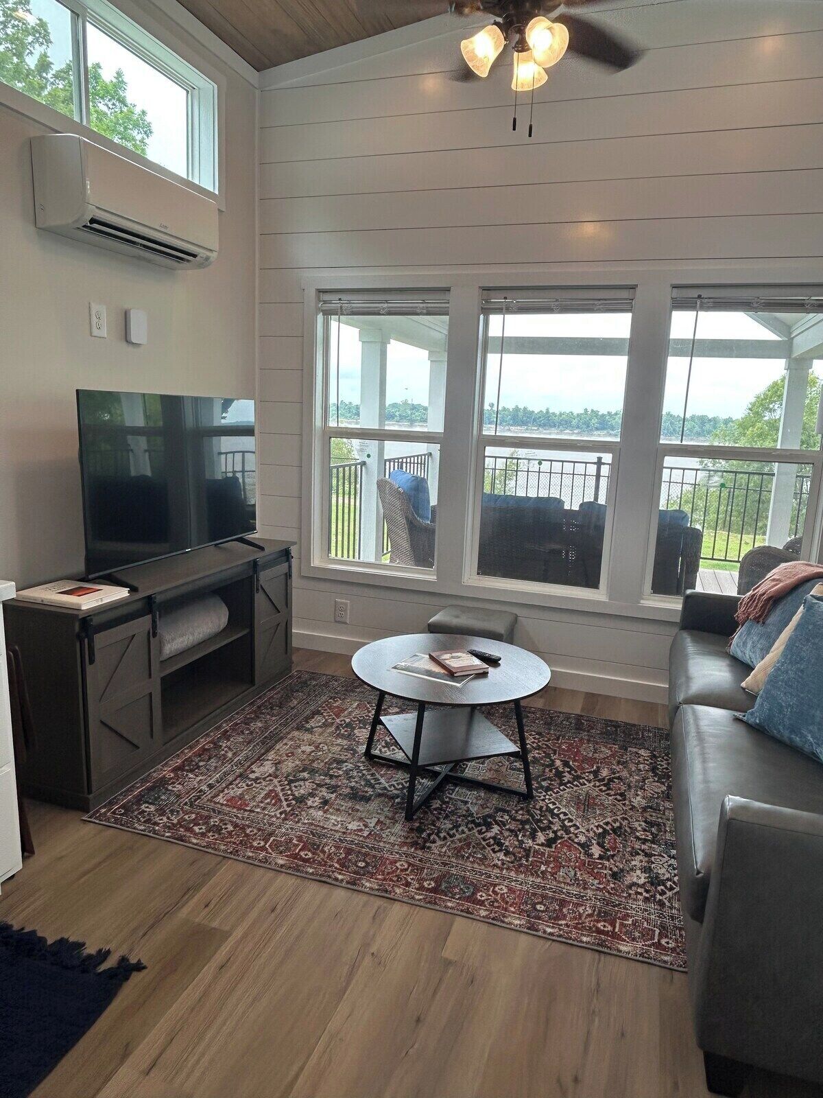 A living room with a television, a round coffee table, and a sofa. There are large windows providing a view of the outdoors.