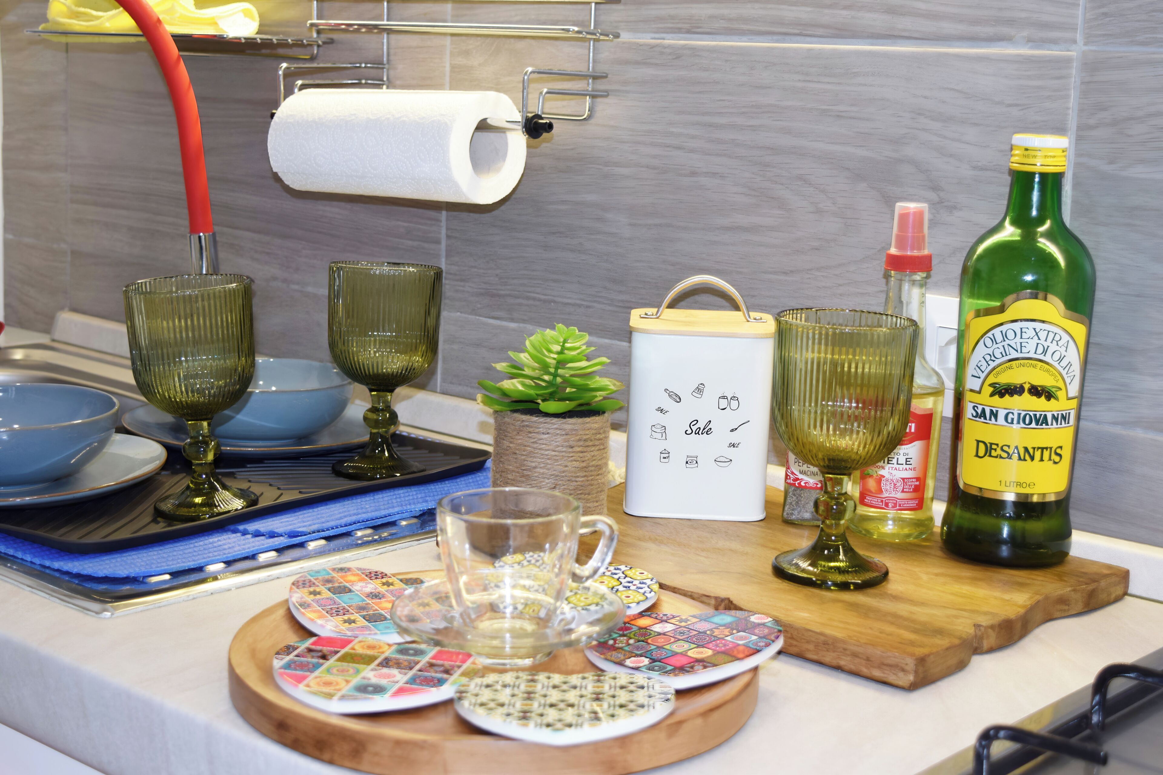 Un comptoir de cuisine sur lequel on trouve divers objets, notamment un rouleau d’essuie-tout, une bouteille verte, un récipient blanc et un plateau avec des sous-verres colorés.