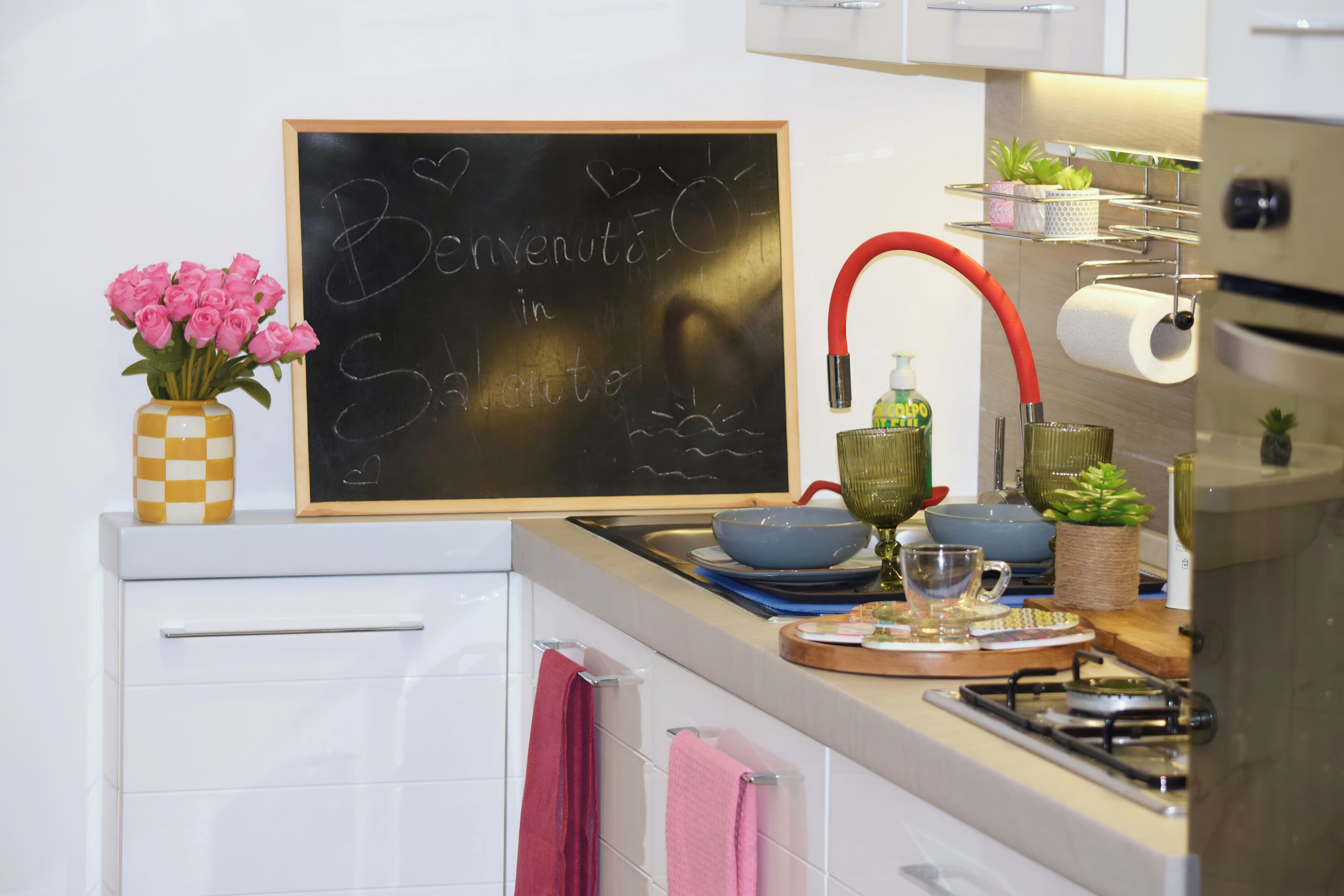 Una cocina con una pizarra mostrando un mensaje, un jarrón de flores rosadas y diversos utensilios de cocina.