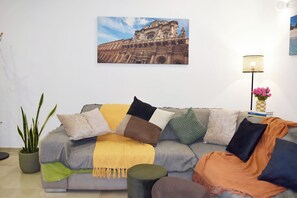 Una sala de estar con sofá, cojines, una planta en maceta, una lámpara y un cuadro en la pared.