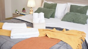 Una cama bien hecha con almohadas y una colcha, una mesita de noche con una lámpara y un libro, y una planta pequeña.