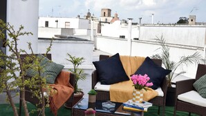 Un espacio de estar en la azotea con mobiliario de mimbre, cojines y una pequeña mesa con libros y un jarrón de flores.