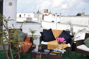 A rooftop seating area with wicker furniture, cushions, and a small table with books and a vase of flowers.