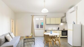 Een knus woongedeelte met een bank, een eettafel met stoelen, een keuken met kastjes, een koelkast en een raam met gordijnen.