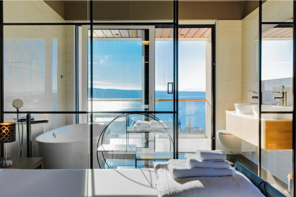 A modern bathroom with a bathtub, toilet, and sink, featuring large windows and a view of the sea.