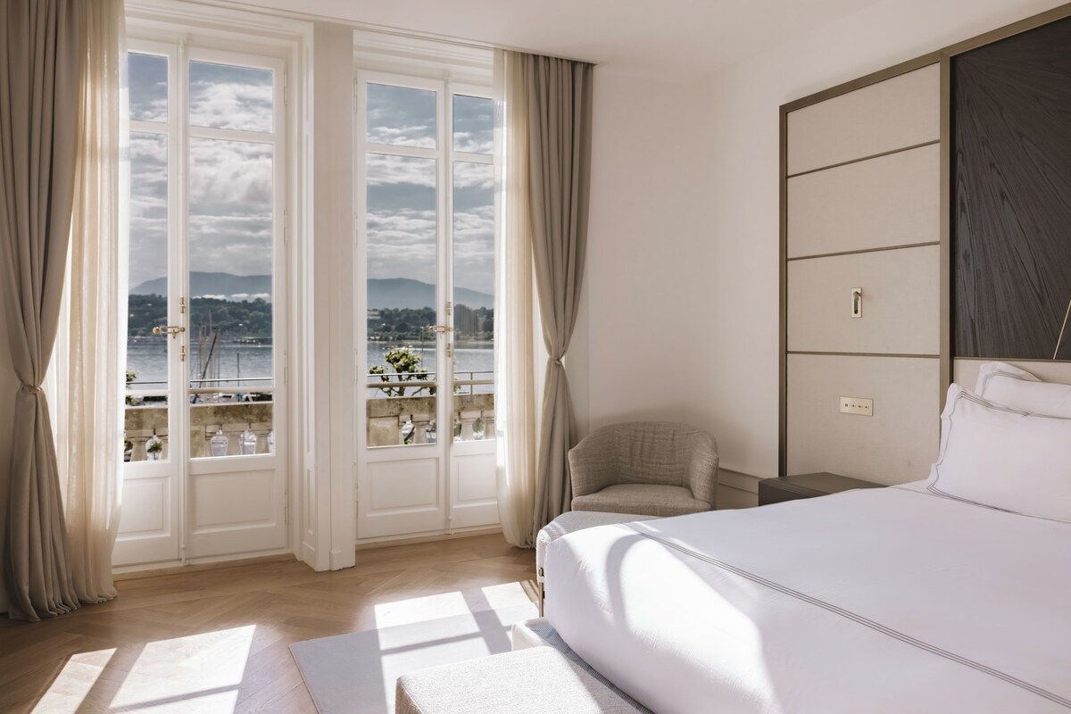 A hotel room with a large bed, a chair, a nightstand, and a view of a body of water through large windows.
