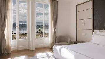 A hotel room with a large bed, a chair, a nightstand, and a view of a body of water through large windows.