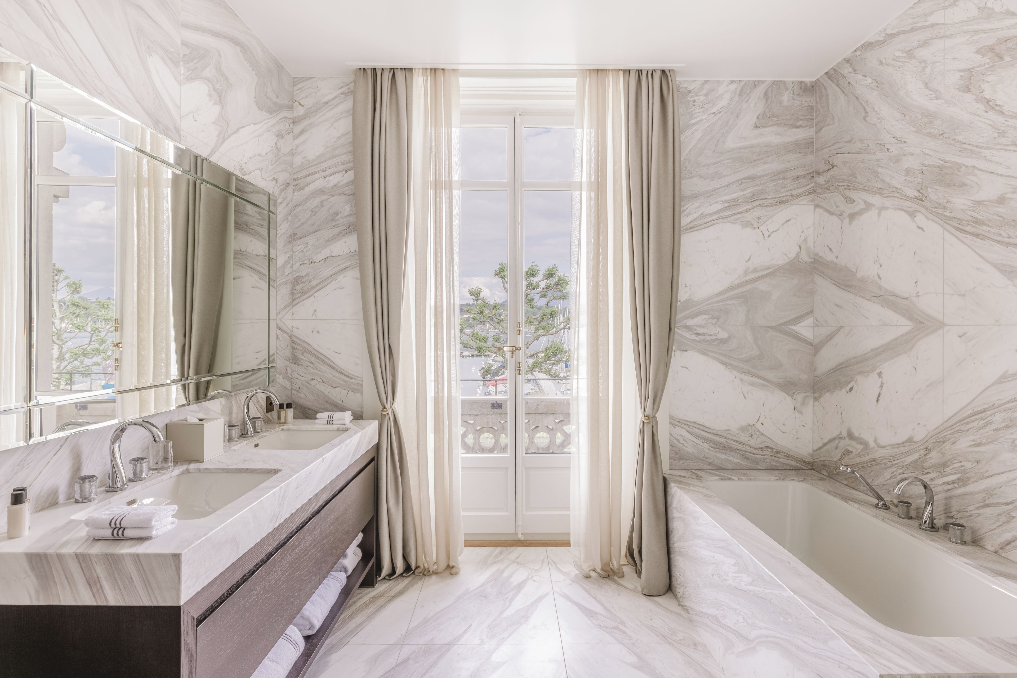 A bathroom with a marble floor, a bathtub, a sink with a mirror, and a view through a glass door.