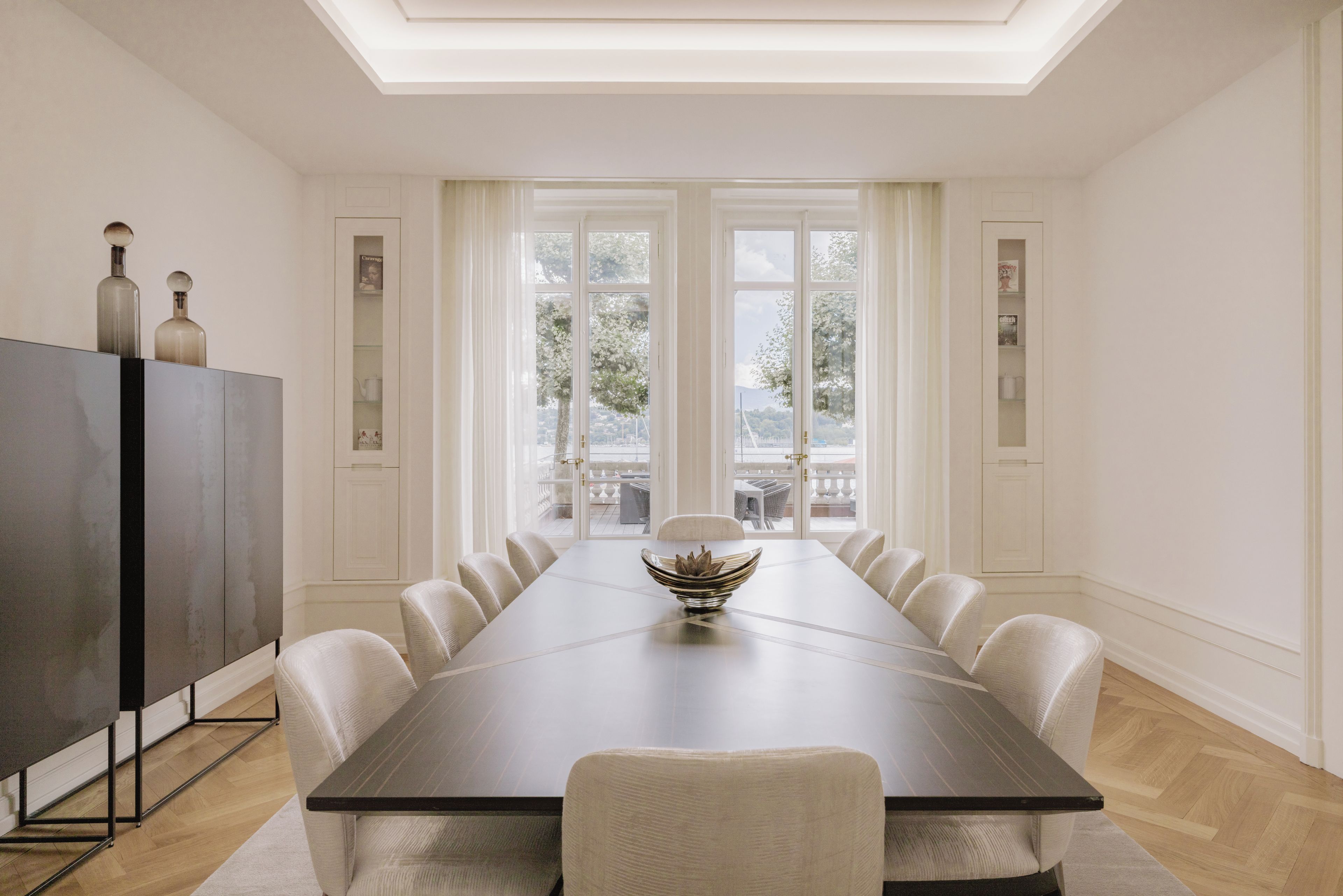 A dining room with a long table, chairs, a modern cabinet, and a view through large windows.