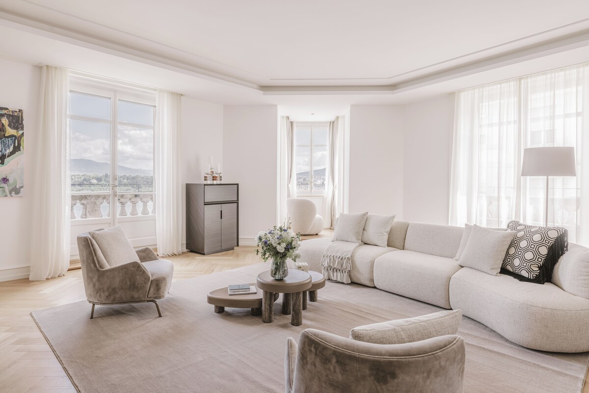 A spacious living room with a large sofa, armchairs, and a coffee table. There is a view of the city through large windows.