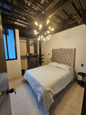 A bedroom with a tufted headboard, a bed with a white quilt, a hanging light fixture, a wardrobe, and a window with blinds.