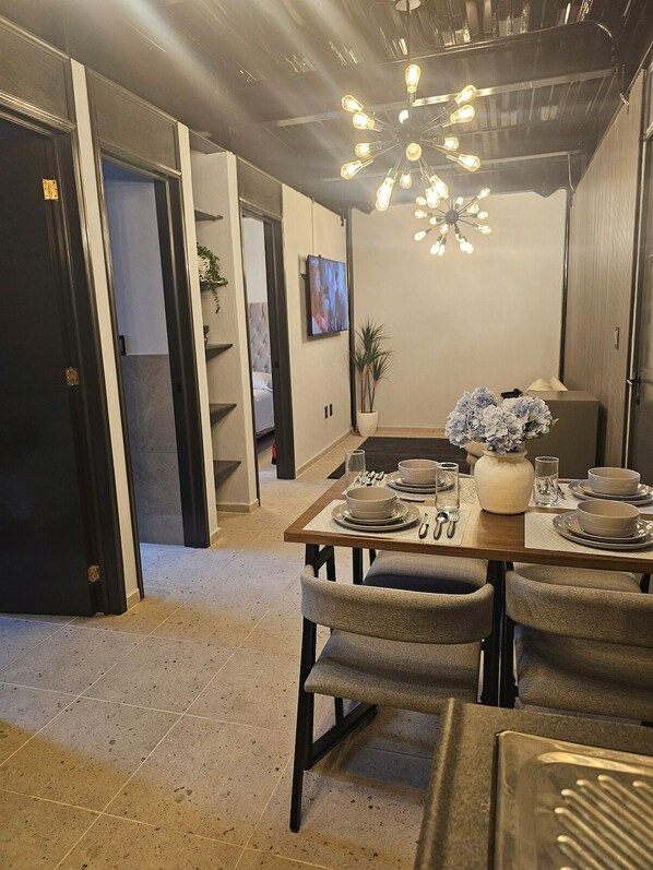 A dining area with a table set for four, a modern chandelier, and a view into a bedroom.