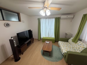 A living room with a flat-screen TV, a ceiling fan, a sofa, a coffee table, and a window with curtains. - Cozy 2-bedroom house in tranquil 渋谷区 with AC (Shibuyaku)