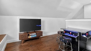 Habitación con televisor de pantalla plana sobre un soporte de madera, una consola de videojuegos y una silla, junto a una pared blanca.
