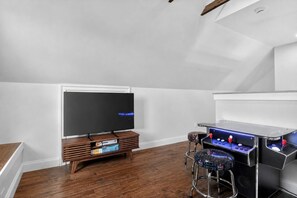 Ein Zimmer mit einem Flachbildfernseher auf einem Holzständer, einer Spielkonsole und einem Sessel, sowie einer weißen Wand.