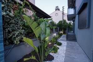 Une ruelle étroite, bordée d’une allée où poussent de petites plantes, et un bâtiment doté d’un toit en pente et d’une fenêtre circulaire.
