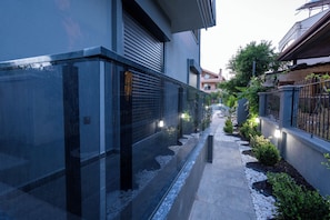 Un estrecho sendero bordeado por edificios con vidrio reflectante y persianas metálicas, que conduce a una entrada con portones.