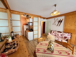 Ein gemütliches Zimmer mit Holzvertäfelung, einem Esstisch mit gemustertem Tischläufer, einer Couch mit dekorativen Kissen und einer Küchenzeile inklusive Kühlschrank und Mikrowelle.