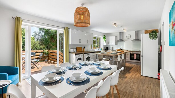 A modern kitchen with white cabinets, a dining area with a white table set for four, and a large window overlooking a garden. - Downs Escape | By My Getaways (Lancing)