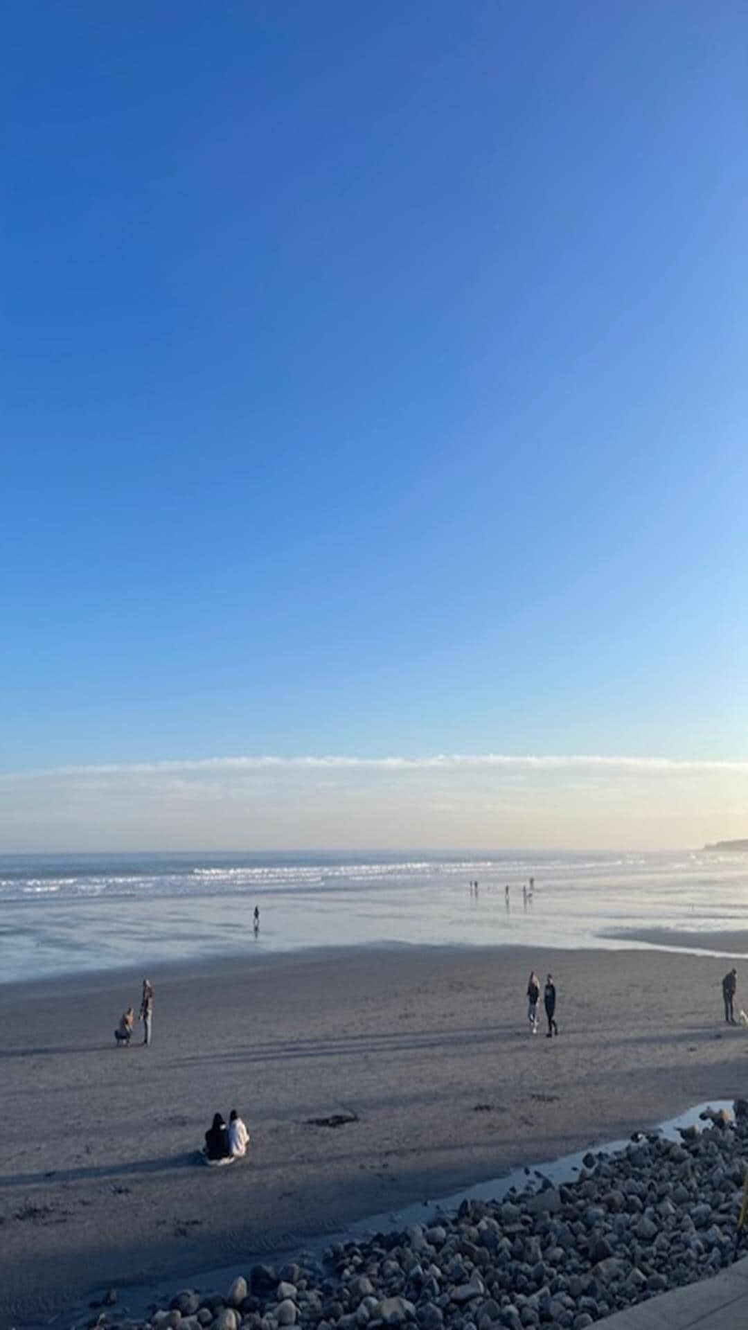 Ein Strand mit Menschen, die auf dem Sand entlanggehen und sitzen, ein klarer Himmel und eine felsige Küste.
