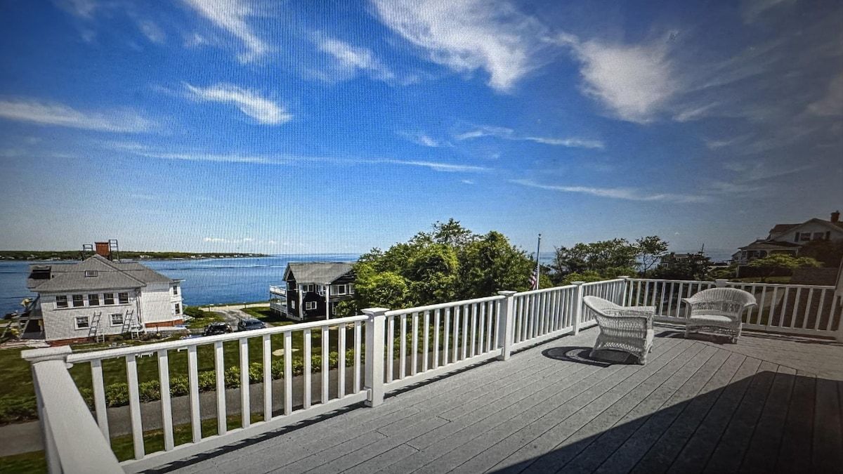 A wooden deck with white railings overlooking a body of water, a grassy area, and a house with a chimney.