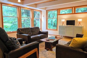 A living room with a sofa, armchair, and coffee table, large windows with a forest view, and a TV on a cabinet. - Private forest escape with creekside trails and firepit (Port Angeles)