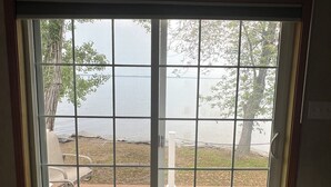 A room with a large sliding glass door leading to an outdoor deck with wooden flooring and a view of a body of water. - Golden Hour Shore House (Swanton)