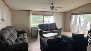 A living room with a sofa, armchairs, a coffee table, a ceiling fan, and a view of the outdoors through large windows. - Golden Hour Shore House (Swanton)