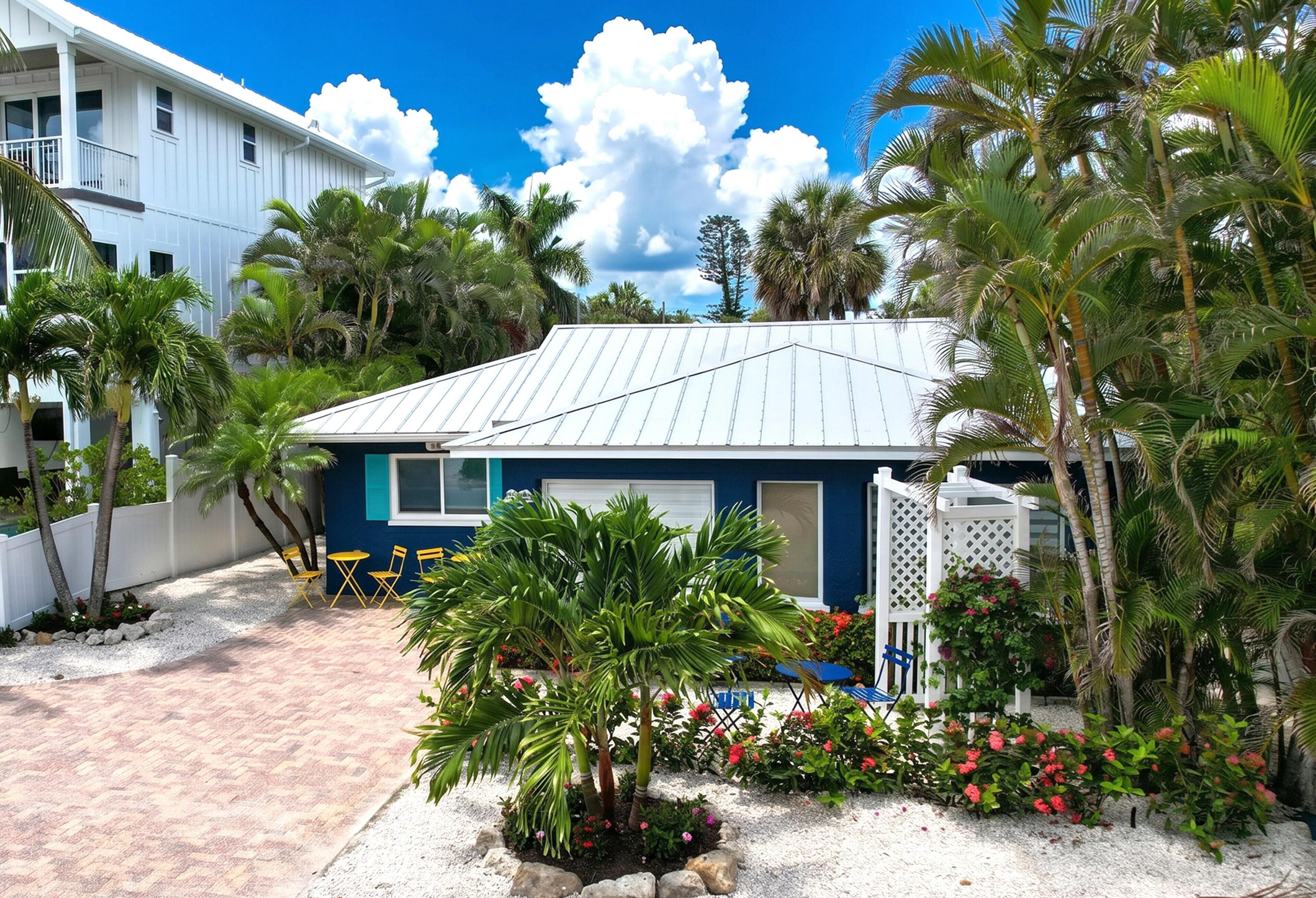A house with a white exterior, a blue door, and a covered patio area with chairs and a small palm tree.