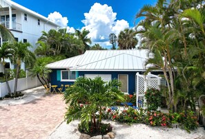 A house with a white exterior, a blue door, and a covered patio area with chairs and a small palm tree.